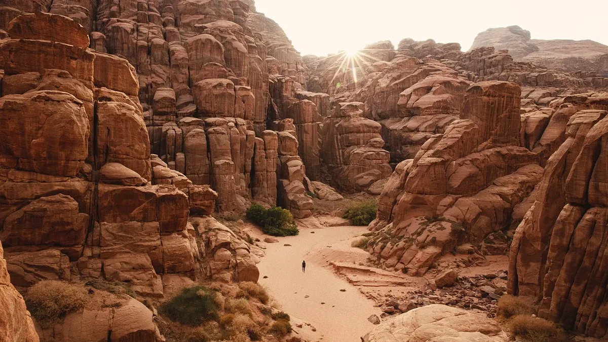 A vast canyon with towering red rock formations, a sandy path, and a person walking in the distance.