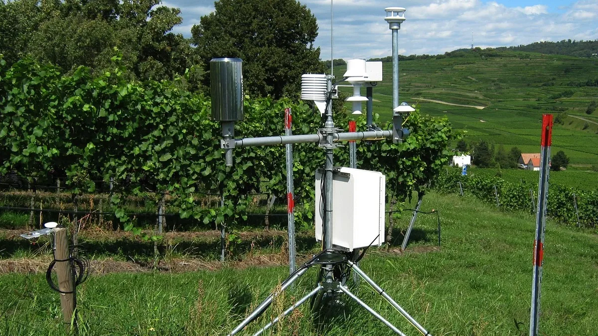 A weather station with various instruments and equipment, including a rain gauge, anemometer, and temperature sensor, set up in a grassy field with trees and hills in the background.